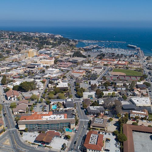 An aerial view of a coastal town with residential and commercial buildings, roads, and a harbor in the background leading to the sea.