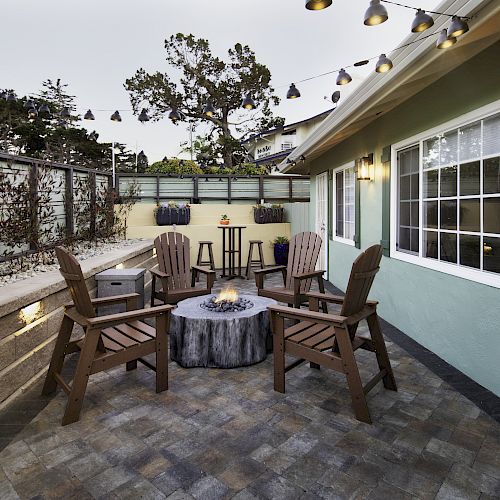 A cozy outdoor patio with a fire pit, four wooden chairs, string lights, a side table, and fenced landscaping in the background.