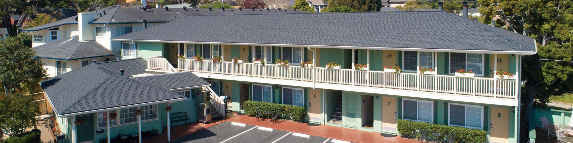 The image shows a two-story motel with a gray roof, green exterior walls, and an empty parking lot in front. Trees and hills are visible in the background.