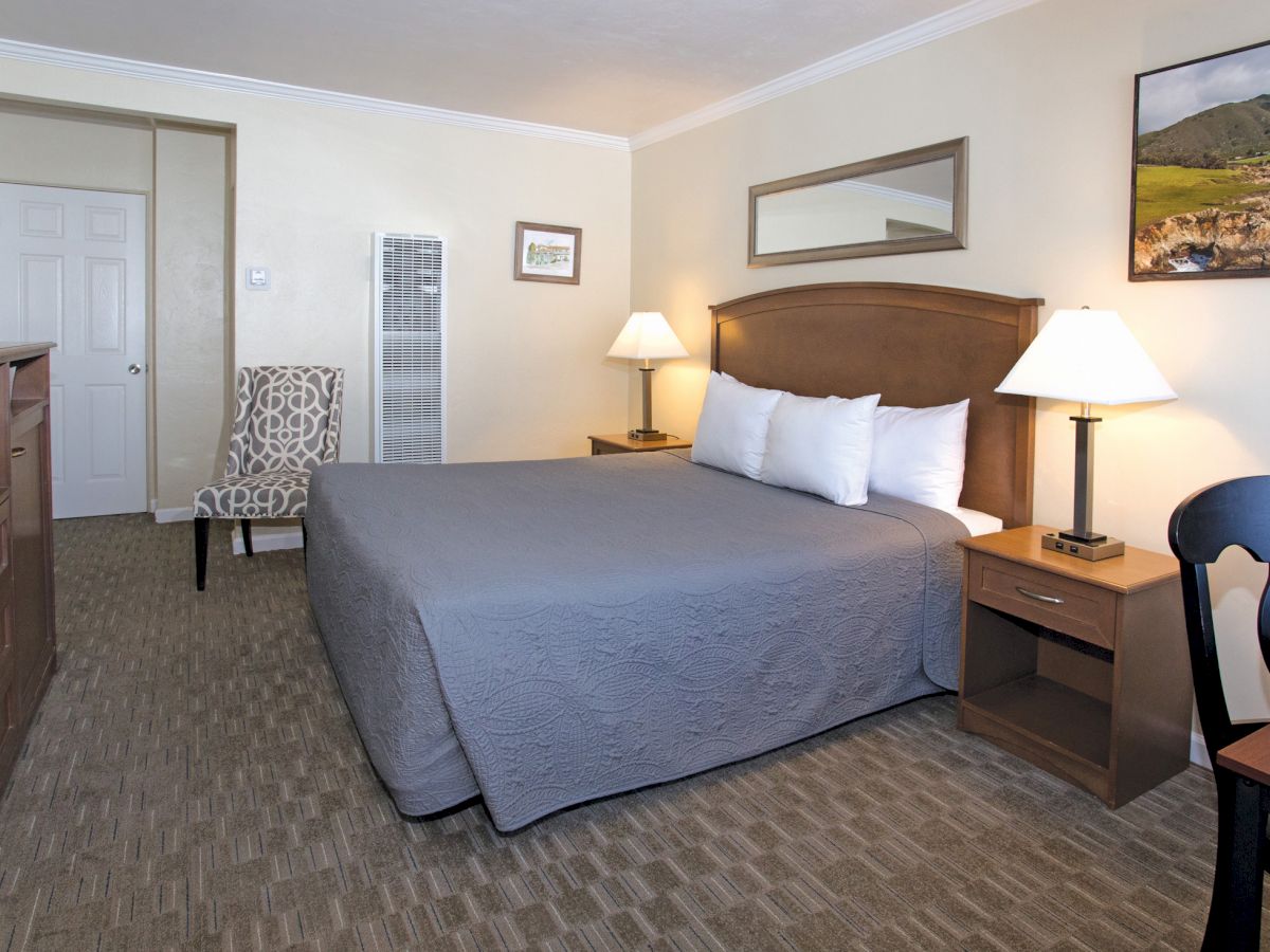 A well-lit hotel room with a large bed, nightstands, lamps, an armchair, dresser, and a framed picture on the wall, in a neat and organized manner.