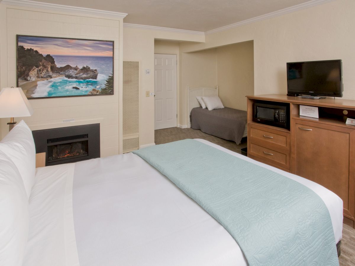 A clean, well-lit hotel room with a bed, fireplace, TV, and a second bed in an adjacent nook. There’s a framed seascape picture on the wall.