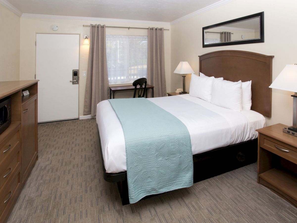 A neatly arranged hotel room with a bed, teal blanket, bedside tables with lamps, a desk by the window, and a TV cabinet with a microwave.