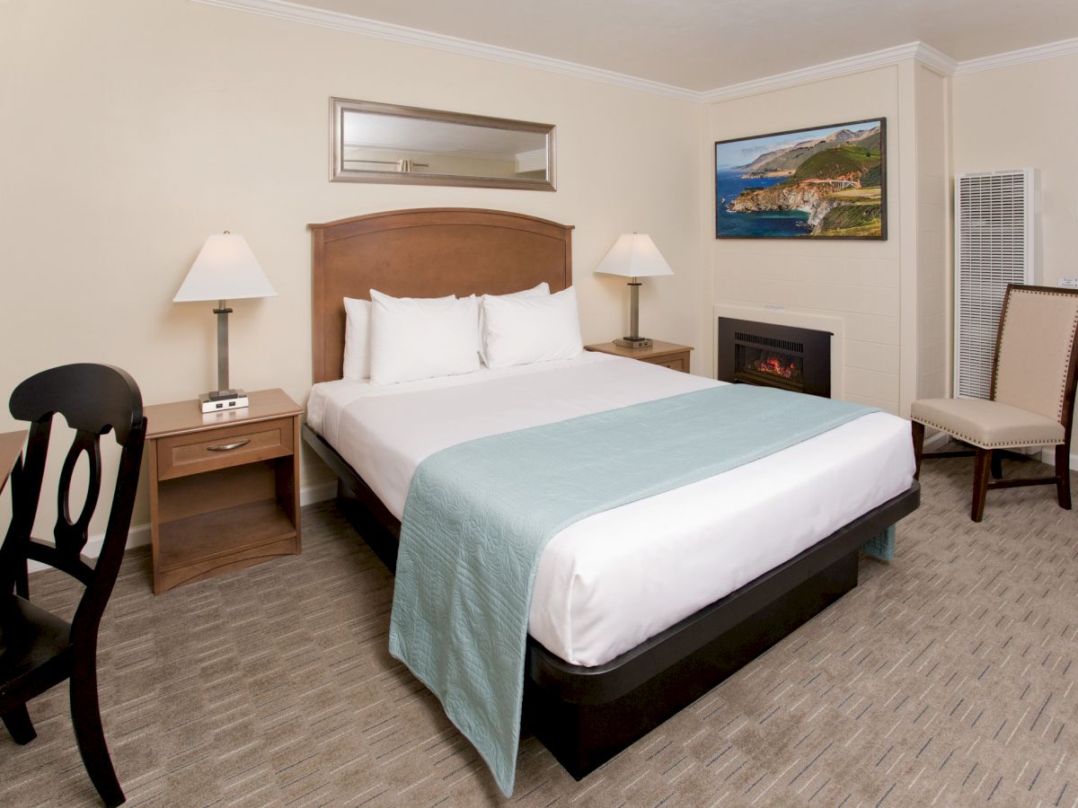 The image shows a neatly arranged hotel room with a king-sized bed, nightstands, a desk, a chair, a fireplace, and wall art.