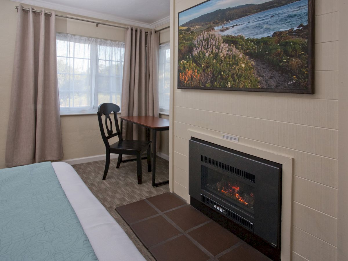 A cozy room with a bed, a small desk with chair, a fireplace, a window with curtains, and a landscape picture on the wall.