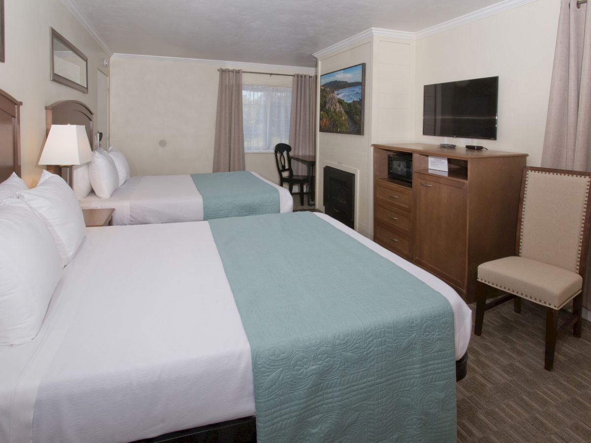 This image shows a hotel room with two beds, a TV, a chair, a window, and a dresser. The room also has a fireplace and neutral decor.