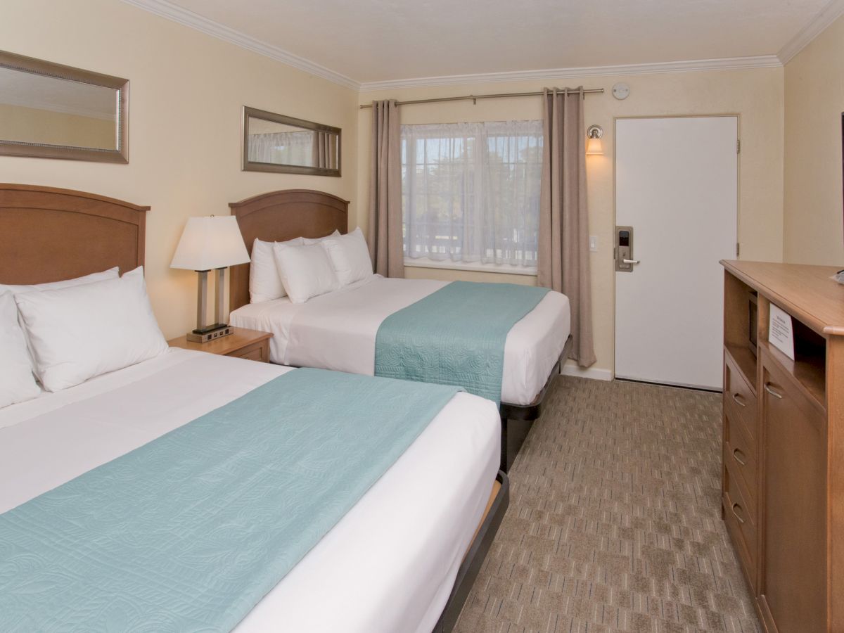 The image shows a tidy hotel room with two double beds, a nightstand with a lamp, a TV on a cabinet, a window with curtains, and a door.