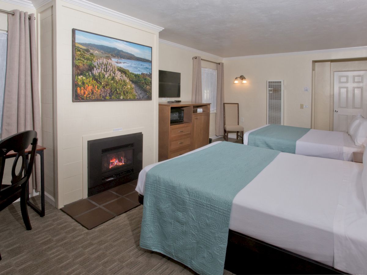 A cozy hotel room with two beds, a fireplace, a flat-screen TV, a desk with a chair, and a scenic picture above the fireplace.