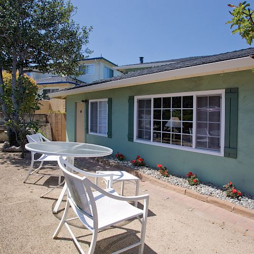 A backyard patio with a round table and chairs on a dirt path, adjacent to a teal house with large windows and minimal landscaping ending the sentence.