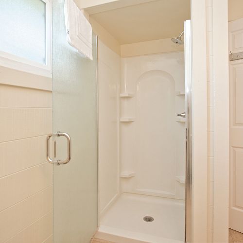 The image shows a clean, modern shower stall with a glass door, white tiles, a towel on the door, a window, and a wall-mounted towel rack.