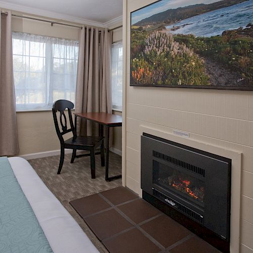 A cozy room with a bed, a fireplace, a wooden table with a chair, curtains on the windows, and a landscape painting above the fireplace.