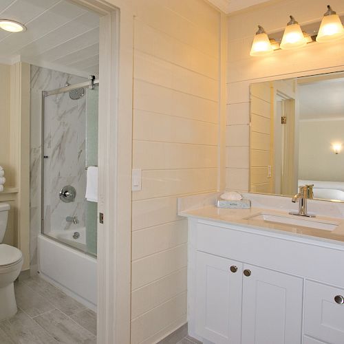 A modern bathroom with a vanity, sink, large mirror, and overhead lights. A bathtub with a shower and toilet are also included in the room.