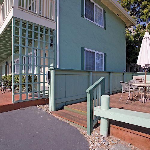 The image shows the exterior of a green building with a balcony and an outdoor seating area, featuring a table, chairs, and an umbrella.