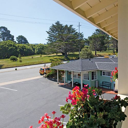 A view from a balcony overlooking a parking lot, small building with flowers in the foreground, and a grassy area with trees in the background.