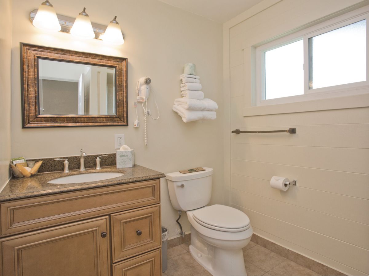 This image shows a bathroom with a vanity, mirror, toilet, window, and towels placed on a shelf above the toilet.