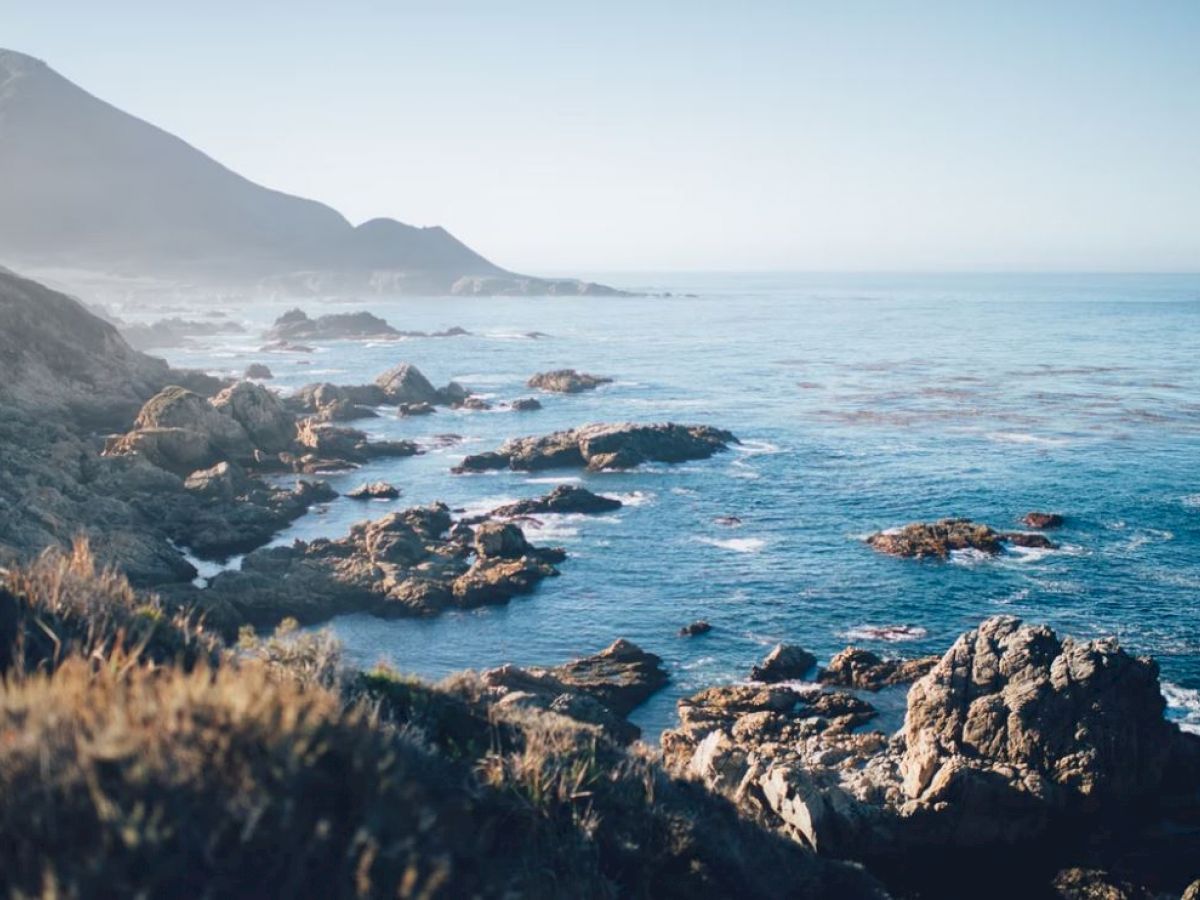 The image shows a scenic coastal view with rugged rocks, blue ocean waters, and distant mountains under a clear sky.