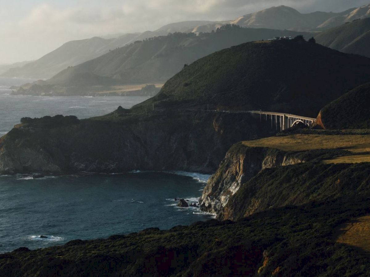 A scenic coastal landscape with rugged cliffs, a curving bridge, and green hills meeting the blue ocean under a partly cloudy sky.