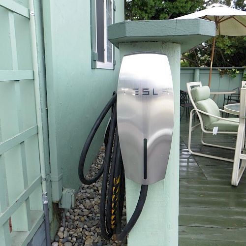 The image shows a Tesla electric vehicle charging station mounted on a post near an outdoor patio area with chairs and a table under an umbrella.