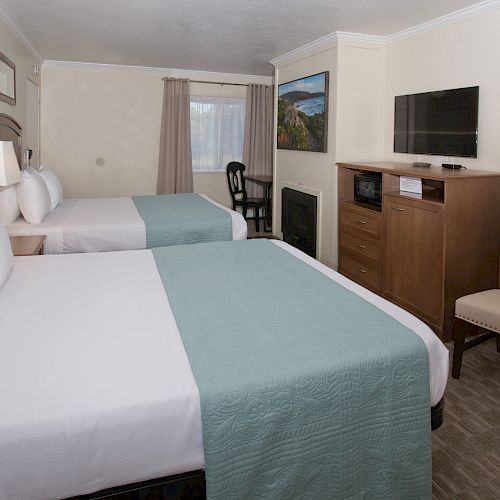 A hotel room with two beds, a TV on a cabinet, a small table with a chair, and light teal bedding. The room also has a window and a lamp.