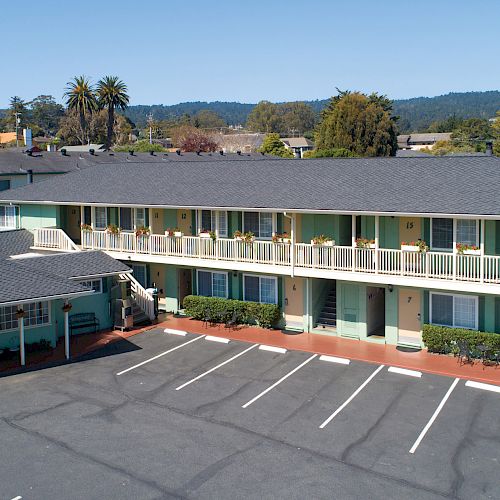 The image shows a two-story motel with a parking area in the front, surrounded by a scenic backdrop of trees and hills.