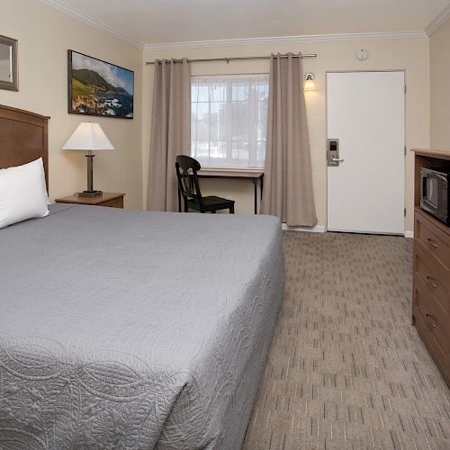 A hotel room with a bed, side table, TV, microwave, desk, chair, and window. The room features minimalistic décor and beige walls.