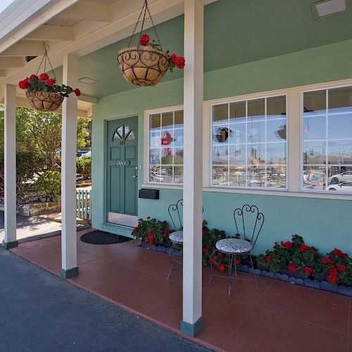 A small porch with hanging plants, a green door, windows, a bench, and potted flowers against a teal building is shown in the image.