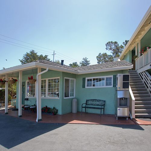 The image shows a small motel or inn with an outdoor stairway, a bench, flower pots, and parking spaces in a bright and sunny setting.