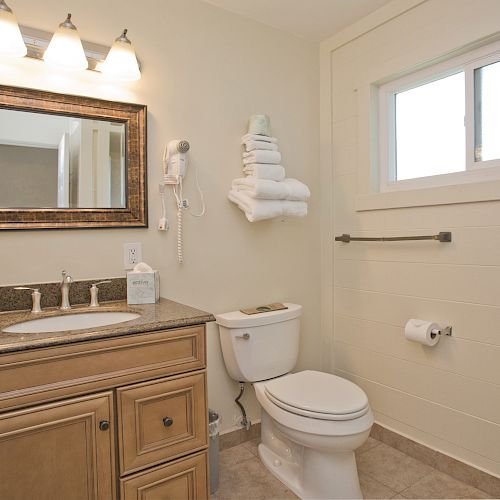 A clean bathroom with a wooden vanity, mirror, sink, and toilet. Towels are stacked on the wall, with a window above providing natural light.