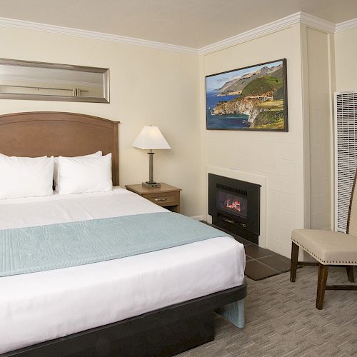 A neatly decorated hotel room with a double bed, two bedside tables with lamps, a fireplace, a chair, and a landscape painting on the wall.