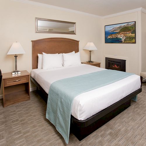 A hotel room with a neatly made bed, two bedside lamps, a desk chair, a straight chair, and wall art above a fireplace.