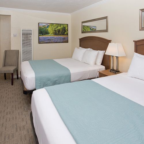 This image shows a hotel room with two queen-sized beds, white bedding, and light blue throws. There are framed pictures and a chair in the corner.