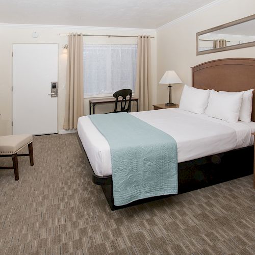 A hotel room with a bed, nightstands, a TV on a dresser, a chair, and a desk in front of a window.