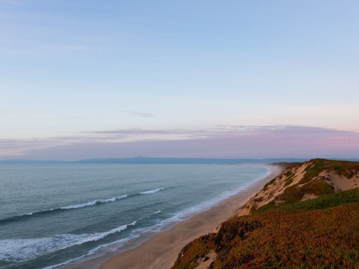 A tranquil coastal scene featuring a sandy beach, rolling waves, and a horizon under a softly lit sky during sunset or sunrise.