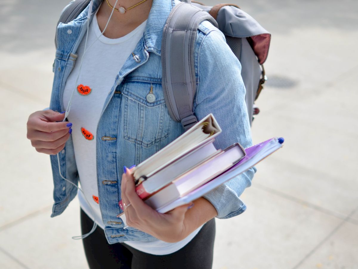 A person wearing a denim jacket and carrying a backpack holds several books and a notebook while walking outside on a sunny day.