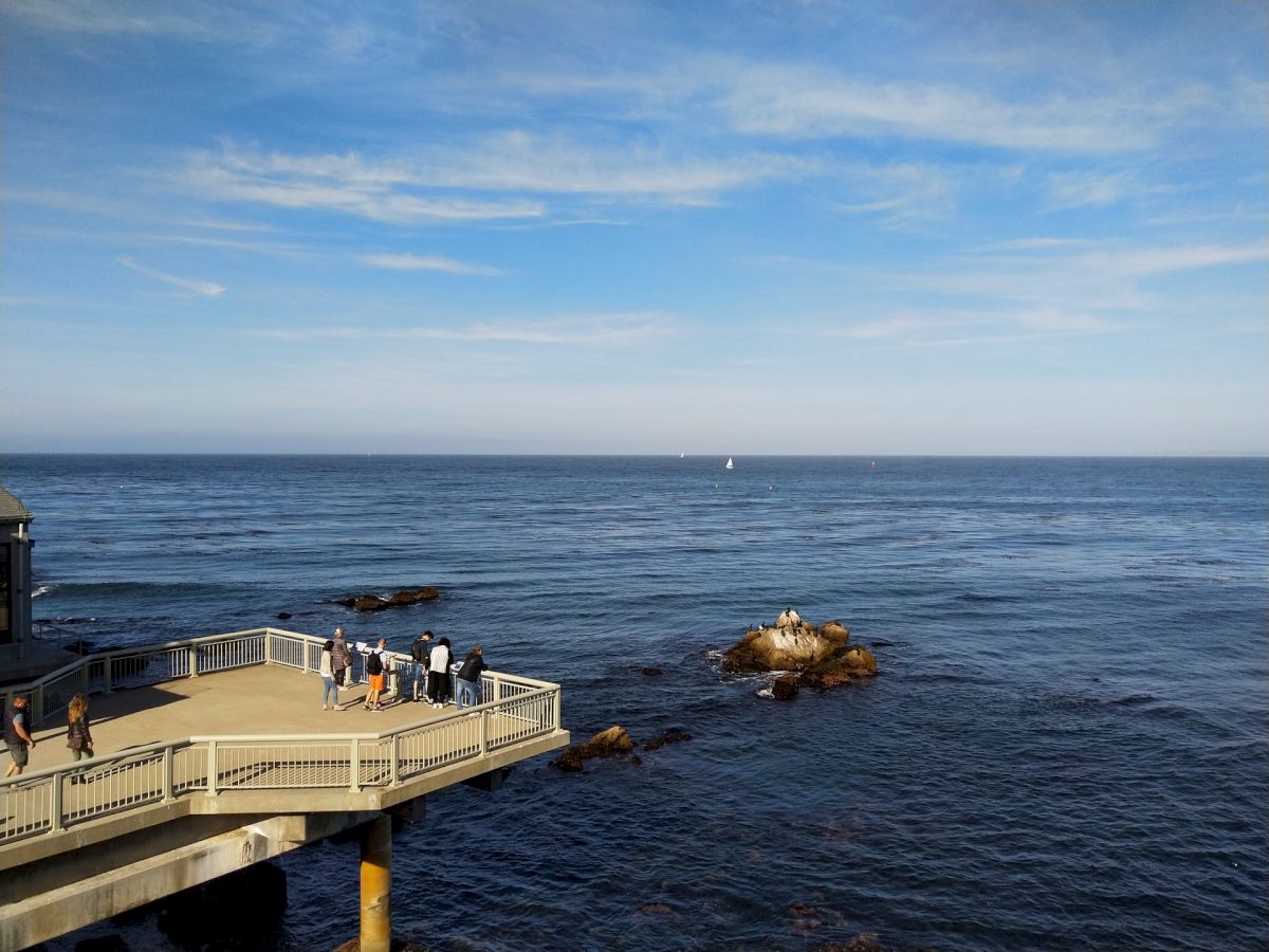 A group of people stands on a seaside observation deck overlooking the ocean, with rocks and calm waves in the background. The sky is partly cloudy.