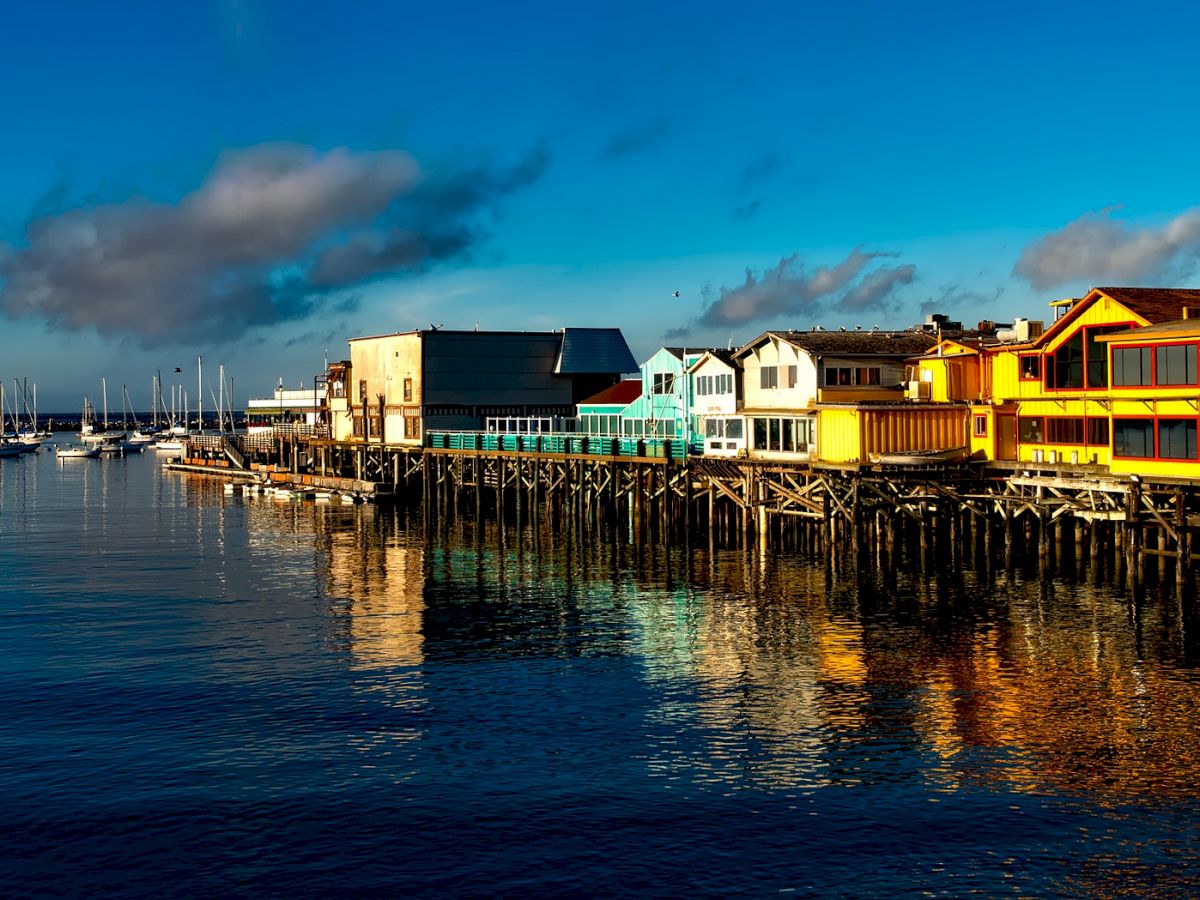 Waterfront scene with colorful buildings on wooden piers, boats docked in the marina, calm water reflecting the structures, and a bright blue sky.