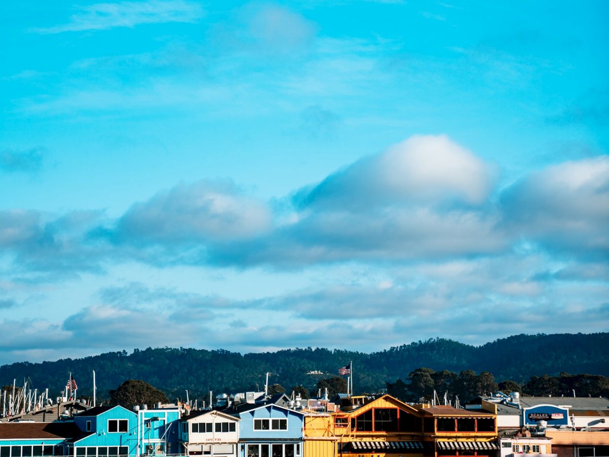 The image shows a waterfront with colorful buildings on a pier, reflecting in the water below, under a sky with fluffy clouds.