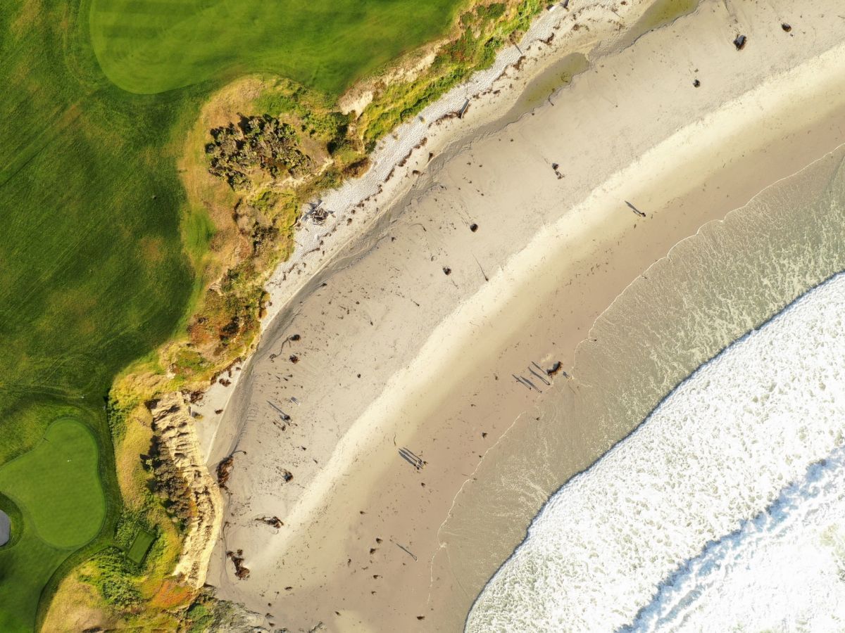 Aerial view of a beach with waves crashing onto the shore, adjacent to a golf course with a green and bunkers visible.