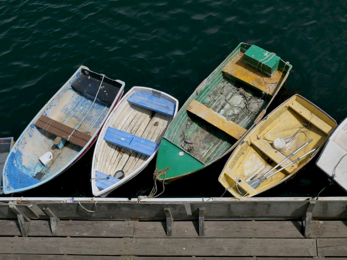 This image shows five small rowboats, each painted differently, moored side by side alongside a wooden dock or pier, floating on calm water.