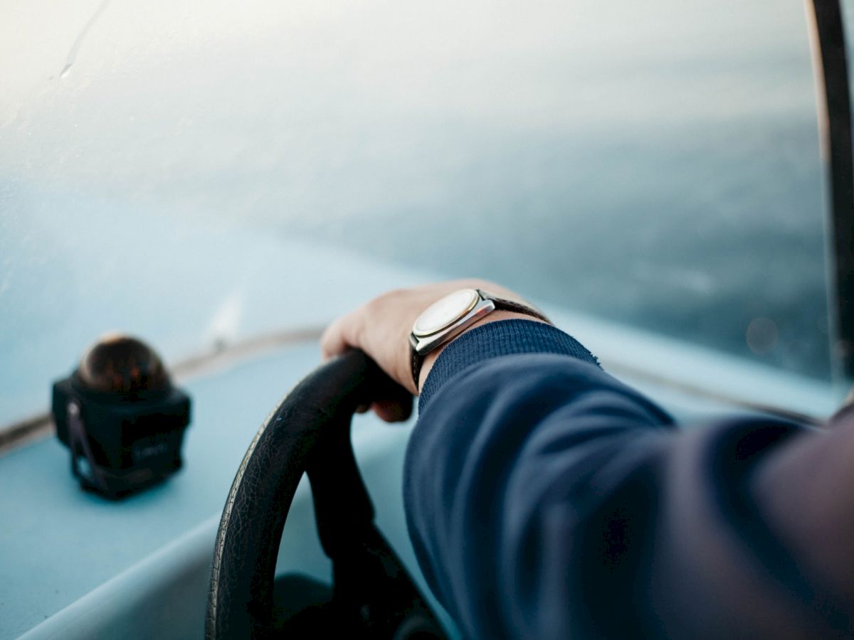 A person wearing a watch is steering a boat, with a small camera or device mounted on the dashboard, and the sea visible ahead.