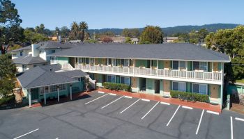 The image shows a two-story motel with a large parking lot in front, surrounded by greenery and distant hills in the background.