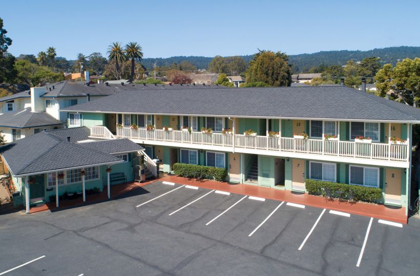 The image shows a two-story motel with a large parking lot in front, surrounded by greenery and distant hills in the background.