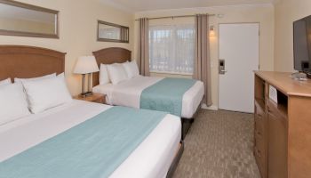 The image shows a hotel room with two double beds, light blue bed runners, a flat-screen TV, a dresser, a lamp, and a window with curtains ending the sentence.