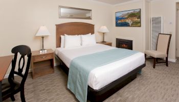 A hotel room with a king-sized bed, two nightstands, lamps, a chair, a desk, and wall art, decorated in neutral and blue tones with a cozy atmosphere.