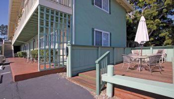 This image shows a light green building with a wooden deck, patio furniture, and an umbrella, adjacent to a parking lot and surrounded by trees.