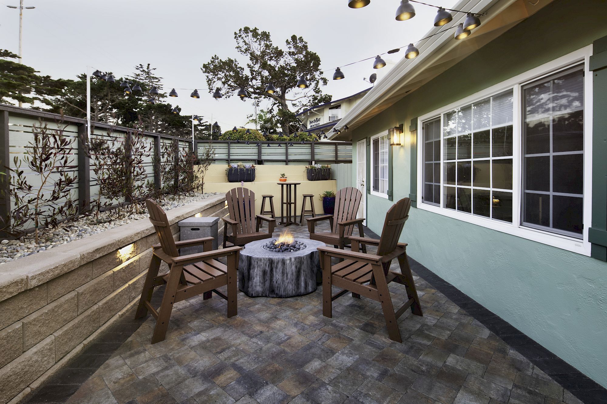 A cozy outdoor patio with wooden chairs around a fire pit, string lights above, and a green house wall with large windows.