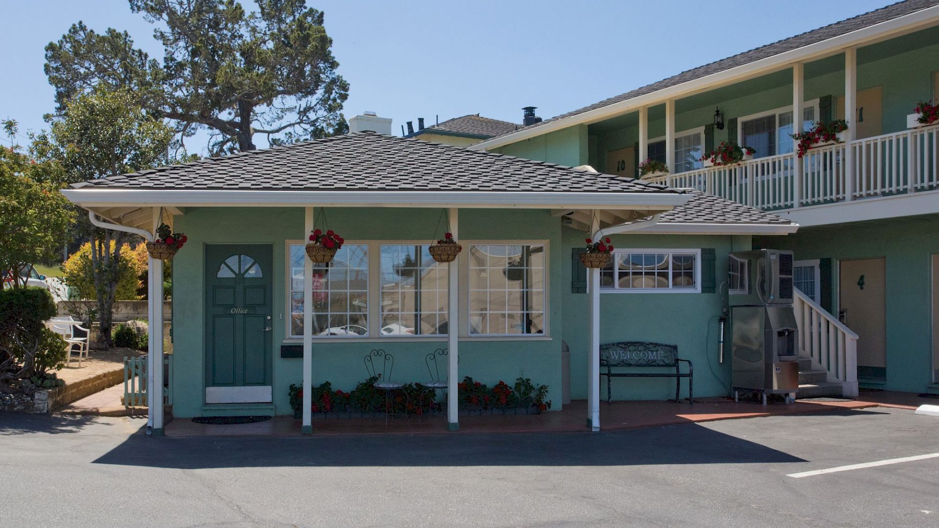 The image shows a turquoise motel with a small porch, potted hanging plants, and a two-story structure behind it, under a clear blue sky.