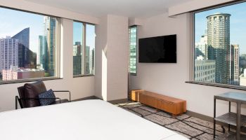 A modern hotel room with large windows offering a city view, a wall-mounted TV, a bed, an armchair, and a small table.