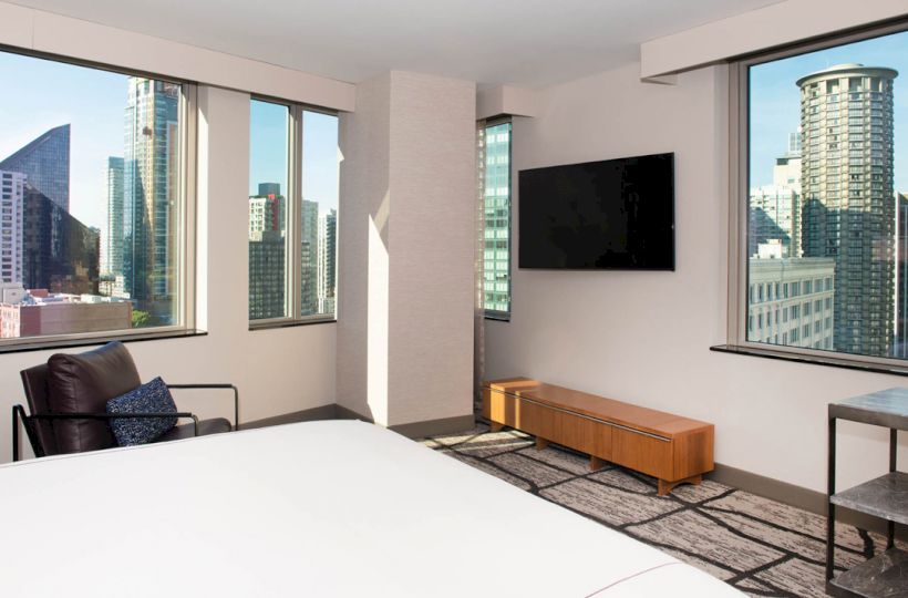 A modern hotel room with large windows offering a city view, a wall-mounted TV, a bed, an armchair, and a small table.