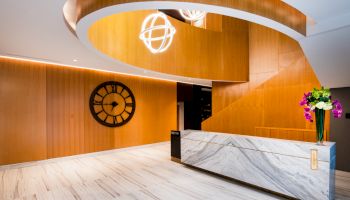 A modern lobby features a marble reception desk, geometric wooden elements, a decorative clock, and contemporary lighting with floral arrangements.