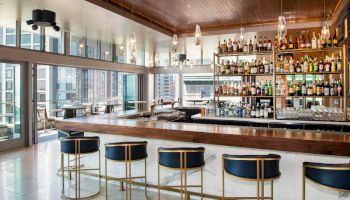 A modern bar with a wooden counter, high stools, shelves filled with assorted bottles, and large windows overlooking a city view, indoors.